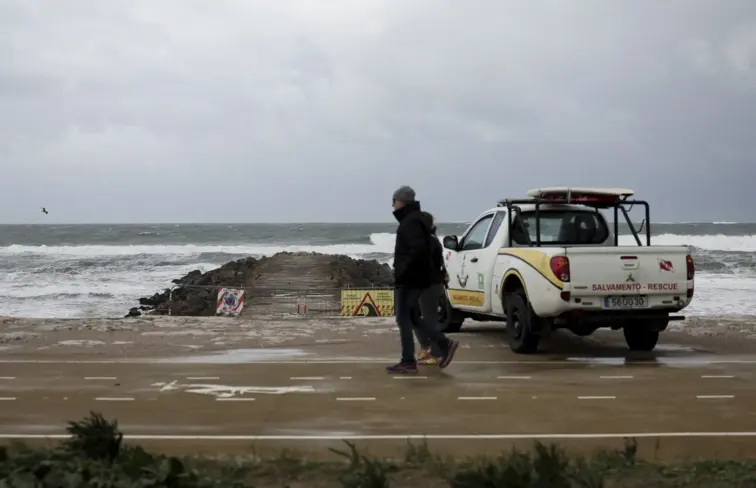 Um carro de salvamento parado perto de um paredão numa praia, num dia cinzento, enquanto uma pessoa com uma capa de chuva passa ao lado.